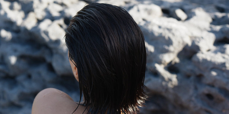 Person mit nassem, dunklem Haar, von hinten gesehen, vor einem felsigen, sonnenbeschienenen Hintergrund stehend. Ihr Gesicht ist nicht sichtbar.