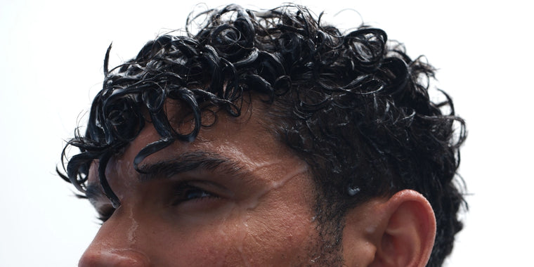 Nahaufnahme einer Person mit nassen, gelockten schwarzen Haaren und Wassertropfen im Gesicht, die vor einem weißen Hintergrund nach oben schaut.