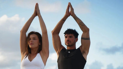 Eine Frau und ein Mann üben am Strand Yoga, indem sie mit über den Kopf gehobenen Armen in Baumhaltung stehen. Die Frau trägt weiße Sportkleidung, der Mann trägt schwarz. Im Hintergrund sind das Meer und ein bewölkter Himmel zu sehen.