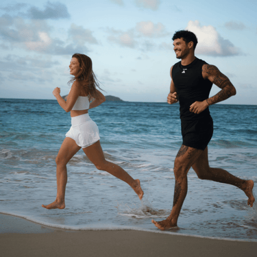 Eine Frau und ein Mann joggen barfuß am Ufer entlang, lächelnd in der Meeresbrise unter einem teilweise bewölkten Himmel. Als Mitglieder des DALUMA HEALTH CLUB PRO MEMBERSHIP kommen sie in den Genuss exklusiver Veranstaltungen zur Förderung von Wohlbefinden und sportlicher Vitalität.