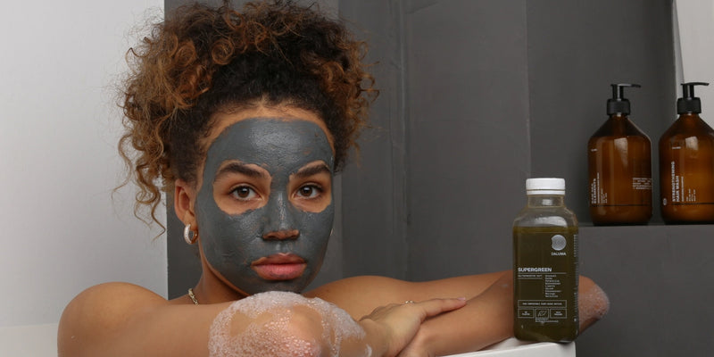 Eine Frau mit einer grauen Gesichtsmaske und lockigem Haar sitzt in einer Badewanne mit Blasen am Arm. Pflegeflaschen und eine grüne Saftflasche stehen neben ihr auf dem Wannenrand.