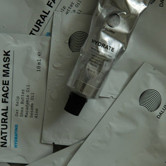 Mehrere DALUMA HYDRATING ESSENTIALS Produkte, darunter eine Metalltube und flache Päckchen mit der Aufschrift Natural Face Mask und Inhaltsstofflisten, sind auf einer wassergetränkten Fläche verstreut. Das DALUMA-Logo erscheint auf jedem Produkt.