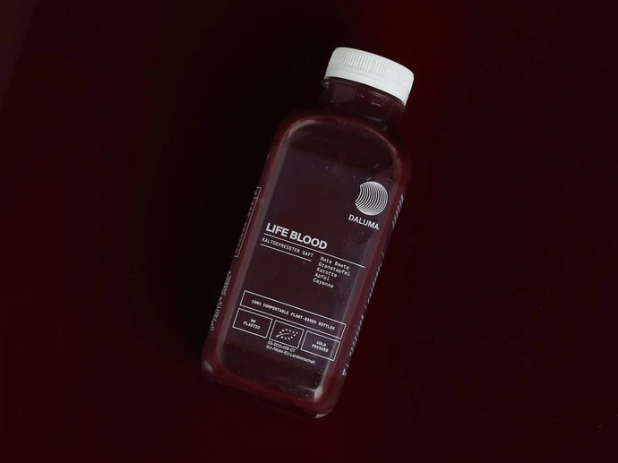 Eine Plastikflasche mit der Aufschrift LIFE BLOOD von DALUMA, die eine tiefrote Flüssigkeit enthält, liegt auf einer dunkelkastanienbraunen Fläche. Auf dem Etikett sind die Inhaltsstoffe angegeben, und die Flasche hat einen weißen Verschluss.
