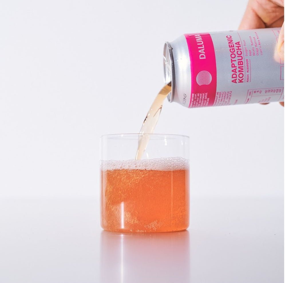 Eine Hand gießt DALUMA MAGIC MUSHROOM DRACHENFRUCHT KOMBUCHA, reich an probiotischen Kulturen, aus einer Dose in ein klares Glas auf einer weißen Oberfläche, um seine Wirkung für die Darmgesundheit hervorzuheben.