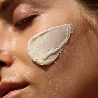 Nahaufnahme des Gesichts einer Person mit geschlossenen Augen, auf der die cremige Gesichtsmaske 3x MASK DUO von DALUMA mit Peelingpartikeln auf der Wange liegt. Die Haut sieht glatt und entspannt aus.