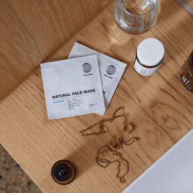 Drei DALUMA 3x Mask Duo Pakete, ein kleines Glas, ein Glas und Goldschmuck sind kunstvoll auf einer Holzfläche neben einem weißen Waschbecken angeordnet.