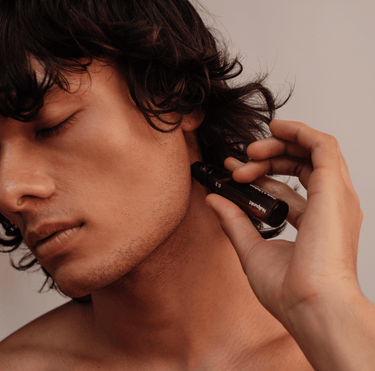 Eine Person mit mittellangem, dunklem Haar trägt vor einem neutralen Hintergrund das Serum COMPLETE COLLECTION von Nullpunkt aus einer Tropfflasche mit geschlossenen Augen sanft im Nacken auf.