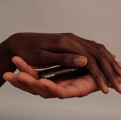 Zwei Hände halten sanft eine kleine, dunkle Flasche aus der NULLPUNKT COMPLETE COLLECTION von Nullpunkt vor einem neutralen Hintergrund, der an Achtsamkeit und Ruhe erinnert.