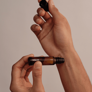 Eine Person hält einen Braunglas-Rollerball-Flakon aus der NULLPUNKT COMPLETE COLLECTION von Nullpunkt in der Hand und nimmt die schwarze Kappe vor einem minimalistischen, neutralen Hintergrund ab.