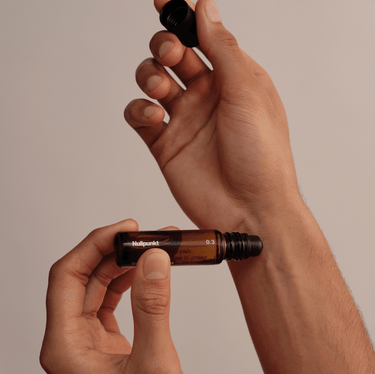 Eine Person hält einen Braunglas-Rollerball-Flakon aus der NULLPUNKT COMPLETE COLLECTION von Nullpunkt in der Hand und nimmt die schwarze Kappe vor einem minimalistischen, neutralen Hintergrund ab.