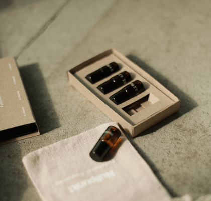 Vier kleine schwarze Fläschchen aus dem Nullpunkt Discovery Set von Nullpunkt sind auf einer strukturierten Oberfläche in einer Pappschachtel angeordnet. Eine Flasche steht außerhalb der Schachtel neben einem hellen Stoffbeutel mit schwacher Aufschrift.