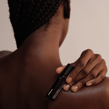 Eine Person mit geflochtenem Haar hält eine glatte NULLPUNKT GRADE 0.1 RELIEVE Hautpflegeflasche von Nullpunkt gegen ihre Schulter, wobei ihre glatte Haut und ihre eleganten Zöpfe vor einem neutralen Hintergrund hervorgehoben werden.