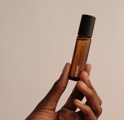 Eine Hand hält eine kleine Braunglas-Tropfflasche mit einer schwarzen Kappe vor einem beigen Hintergrund. Auf dem Etikett steht 