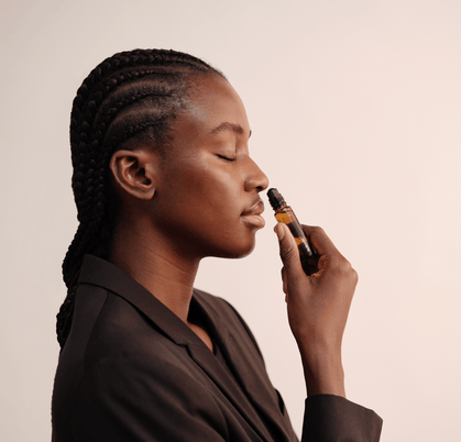 Eine Frau mit geflochtenem Haar und geschlossenen Augen hält einen Nullpunkt GRADE 0.1 RELIEVE Aromatherapie-Ölroller an ihre Nase, als ob sie daran riechen würde, während sie einen schwarzen Blazer vor einem hellen Hintergrund trägt.