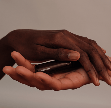 Eine Nahaufnahme von zwei Händen, einer dunkleren und einer helleren, die sanft eine kleine schwarze NULLPUNKT GRADE 0.1 RELIEVE Flasche von Nullpunkt vor einem minimalistischen, neutralen Hintergrund halten.