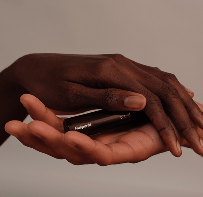 Eine Nahaufnahme von zwei Händen, einer dunkleren und einer helleren, die sanft eine kleine schwarze NULLPUNKT GRADE 0.1 RELIEVE Flasche von Nullpunkt vor einem minimalistischen, neutralen Hintergrund halten.