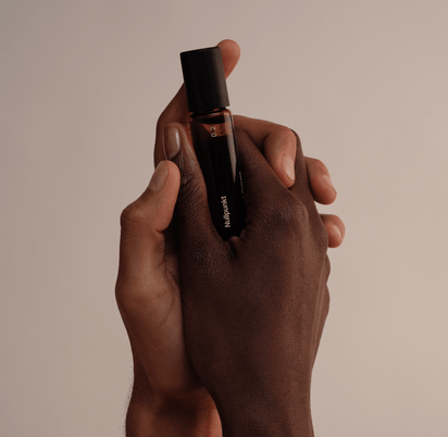 Zwei Hände - eine hellere und eine dunklere - halten sanft den NULLPUNKT GRADE 0.2 RELEASE, einen Parfümflakon aus Braunglas mit schwarzem Verschluss von Nullpunkt, vor einem neutralen beigen Hintergrund.