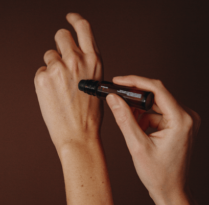 Eine Person rollt eine dunkle Glasflasche mit NULLPUNKT GRADE 0.2 RELEASE von Nullpunkt auf ihrem linken Handrücken vor einem braunen Hintergrund, der die Anwendung dieses Produkts andeutet.