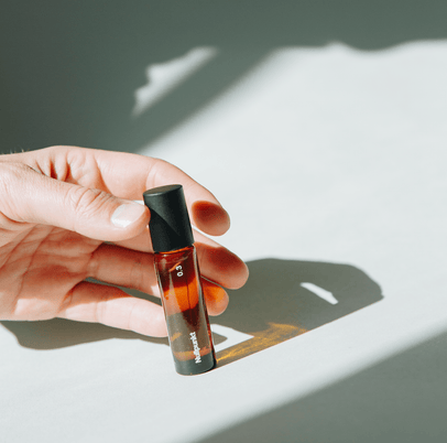 Eine Hand hält einen Flakon NULLPUNKT GRADE 0.3 REFOCUS von Nullpunkt aus Braunglas mit schwarzem Verschluss, der im hellen Sonnenlicht einen scharfen Schatten auf eine weiße Oberfläche wirft.