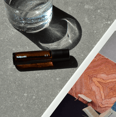 Ein kleines Braunglasfläschchen mit der Aufschrift NULLPUNKT GRADE 0.3 REFOCUS von Nullpunkt steht auf einer grauen Fläche neben einem halbvollen Glas Wasser und einer bunten Magazinseite mit abstrakten Mustern und einem beigen Stuhl.