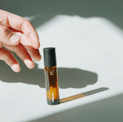 Eine Hand greift nach einer kleinen Braunglasflasche mit schwarzem Verschluss, die die Aufschrift NULLPUNKT GRADE 0.4 REFUEL by Nullpunkt trägt und bei starkem Sonnenlicht einen deutlichen Schatten auf eine helle Fläche wirft.