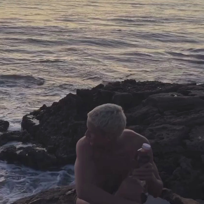 Eine Person mit kurzen, hellen Haaren sitzt bei Sonnenuntergang auf einem Felsen im Meer, hält eine Flasche Daluma BETTER ENERGY SAFTKUR 3 TAGE in der Hand und blickt auf die Wellen, die im Hintergrund schlagen.