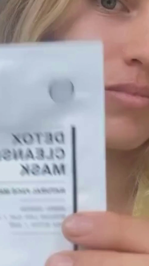 Eine Frau mit langen blonden Haaren hält eine Packung mit der Aufschrift Detox Cleansing Mask in die Kamera, wobei ihr Gesicht teilweise verdeckt wird. Sie scheint in einem Haus zu sein und trägt ein helles Oberteil.