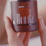Eine Hand hält einen braunen, gefrosteten Tiegel mit der Aufschrift Prebiotic Purifying Cream, wobei der Text teilweise sichtbar ist; der Hintergrund ist leicht unscharf.