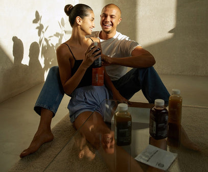 Ein lächelndes Paar sitzt auf dem Boden im Sonnenlicht, hält Getränkeflaschen in der Hand und sieht sich an. Drei weitere Flaschen und Päckchen stehen auf einem Glastisch vor ihnen. Sie sind entspannt und genießen die Gesellschaft des anderen.