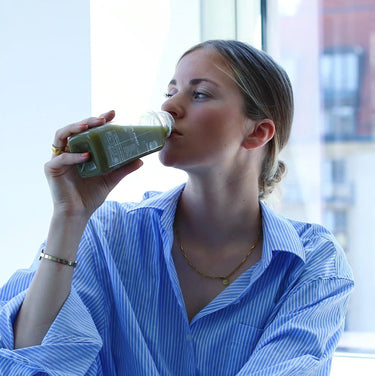 Eine Frau in einem blau gestreiften Hemd genießt DALUMA GREEN JUICE PLUS SAFTKUR 5 TAGE aus einer Glasflasche am Fenster und lässt sich die frischen Bio-Gemüsesäfte schmecken.