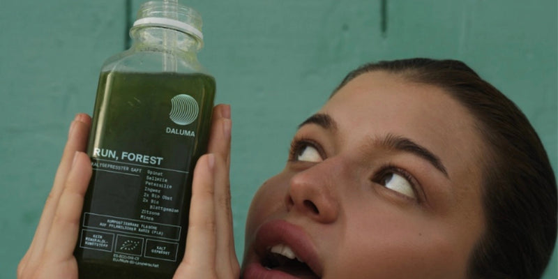 Eine Person mit geschürzten Lippen blickt auf eine durchsichtige Flasche grünen Saftes mit der Aufschrift RUN, FOREST von DALUMA, die sie dicht vor ihr Gesicht hält, vor einem blaugrünen Hintergrund.